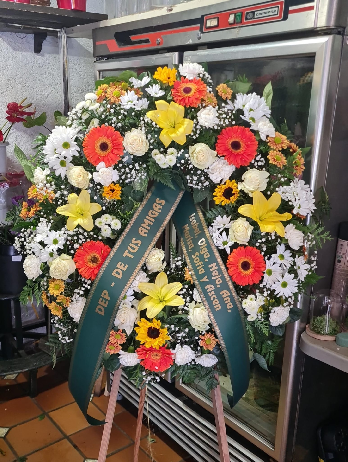 Corona funeraria con gerberas, lilium y margaritas en tonos cálidos